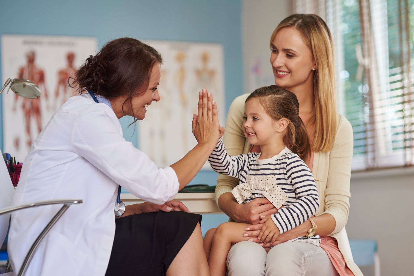 A female doctor in a white coat gives a high-five to a smiling young girl sitting on her mother's lap in a bright clinic, conveying a friendly atmosphere.