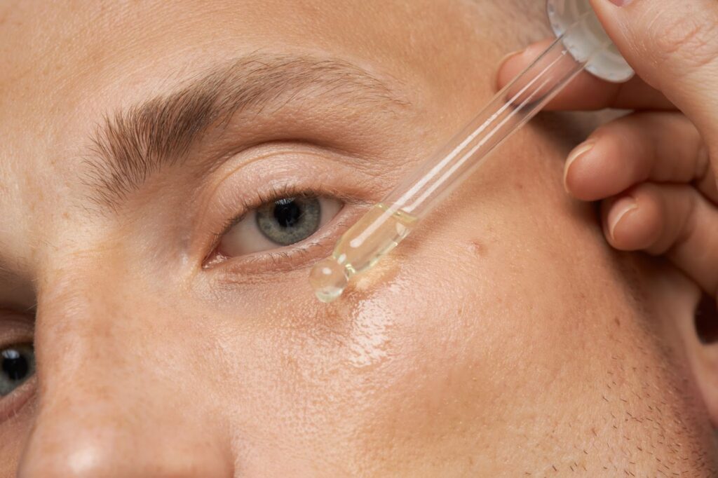 A close-up of a person applying a dropper of liquid serum to their face near the eye.