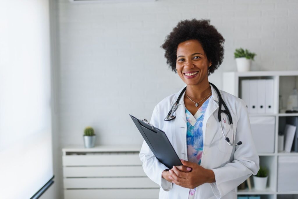 A smiling doctor in a white coat holding a clipboard in a clean office setting.