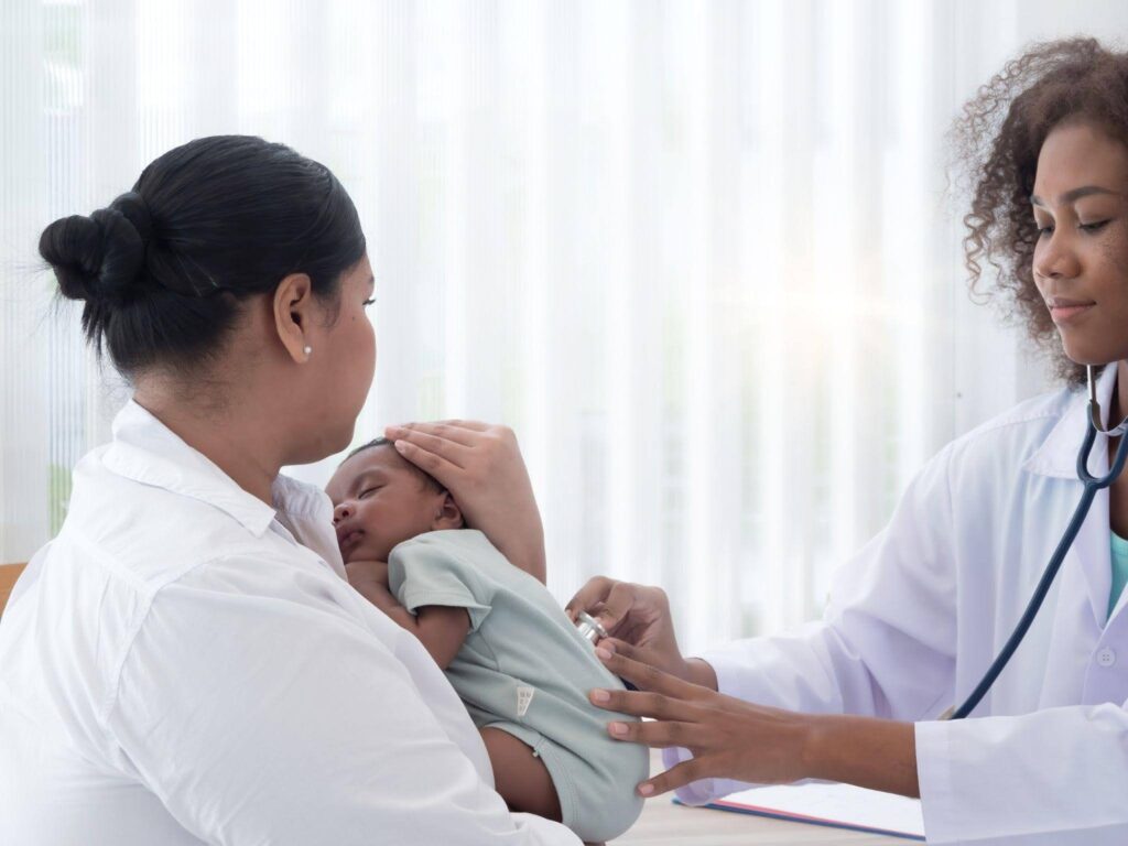 A woman holds a baby while a doctor conducts a medical examination in a clinical setting
