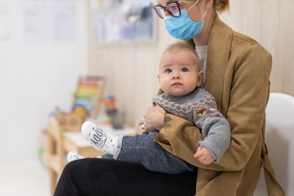 A woman with a face mask holds a baby both demonstrating adherence to health guidelines