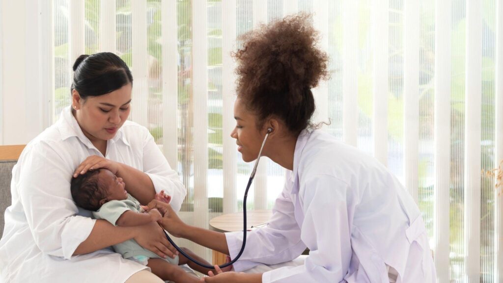 A woman holds a baby while a doctor listens to her heart with a stethoscope in a medical setting