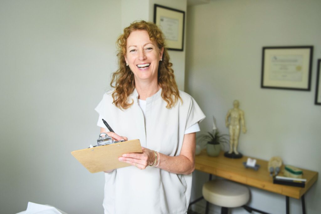 Smiling female healthcare practitioner holding a clipboard and pen in a modern clinic office.